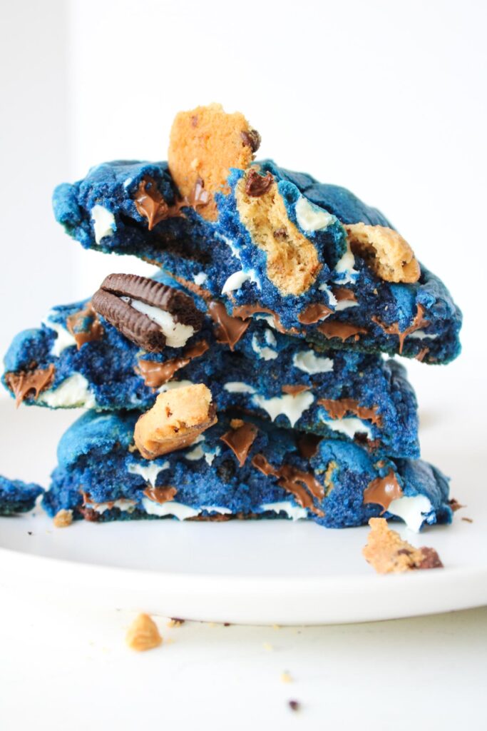 A stack of vibrant blue cookies filled with chocolate chips, crushed Oreos, and pieces of Chips Ahoy, on a white plate.