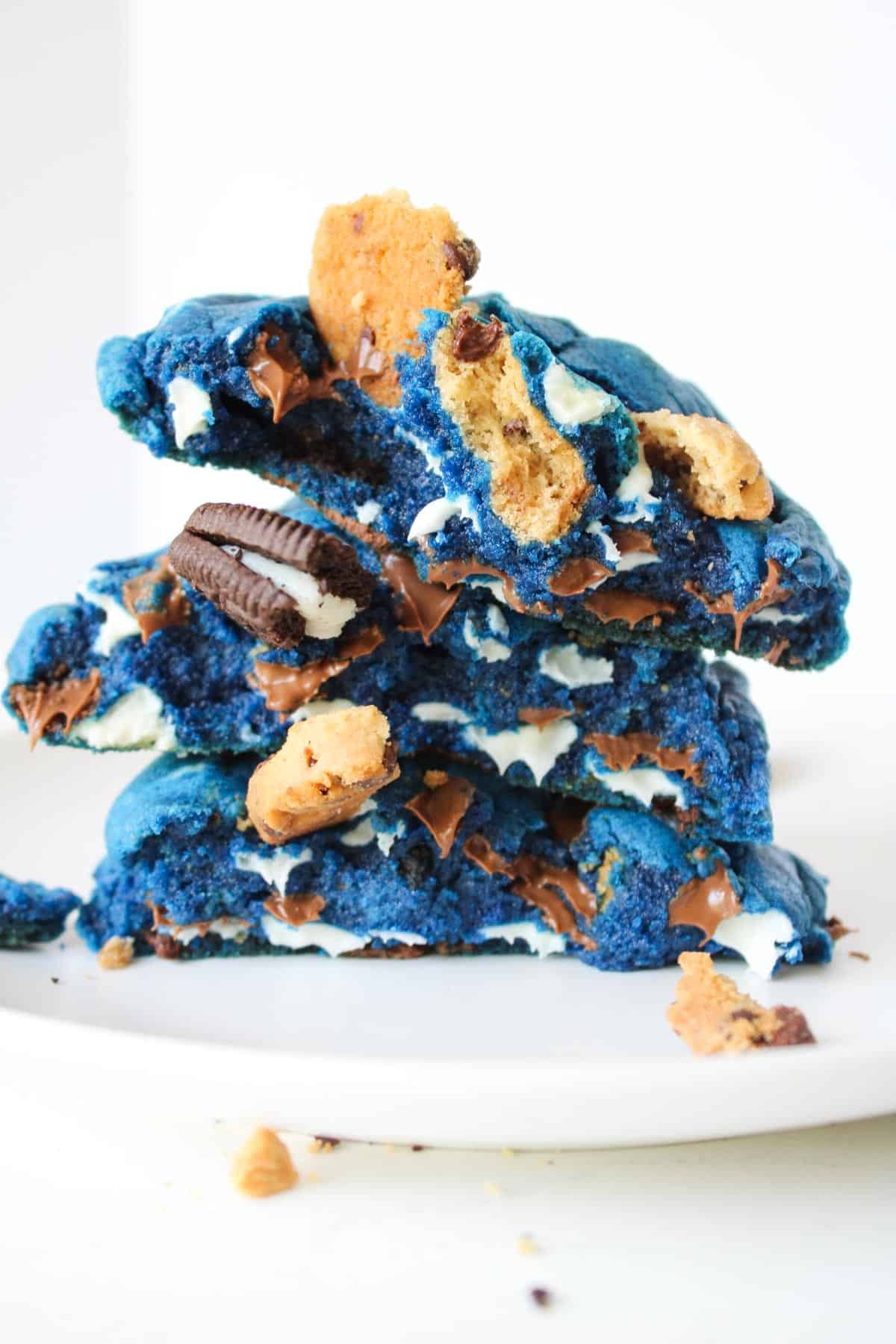 A stack of vibrant blue cookies filled with chocolate chips, crushed Oreos, and pieces of Chips Ahoy, on a white plate.