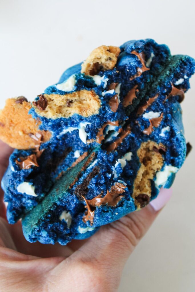 A person holds a vibrant blue cookie filled with chocolate chips and cookie pieces, showcasing its gooey, colorful interior.