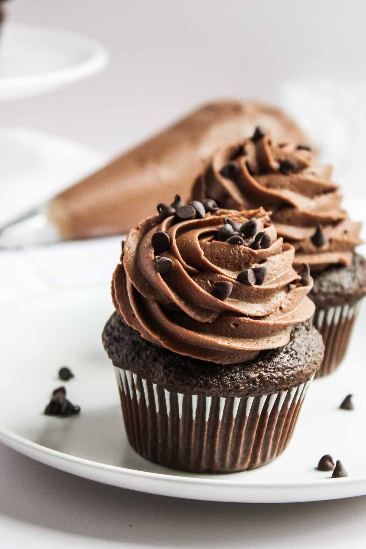 Chocolate cupcakes topped with swirls of chocolate buttercream frosting and mini chocolate chips, with a piping bag in the background.