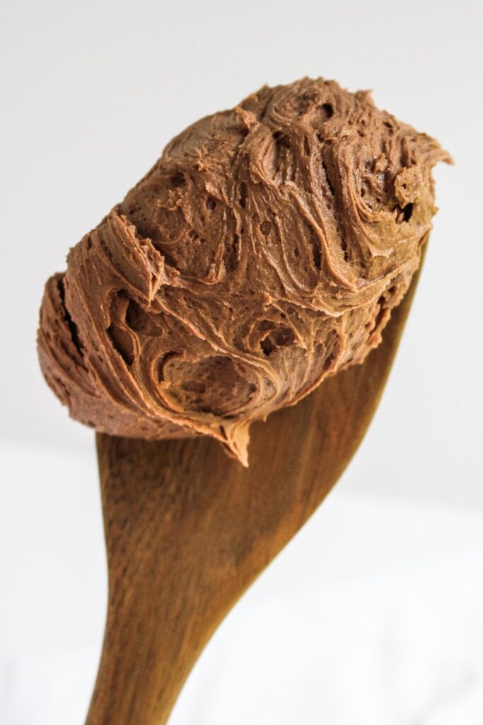 A close-up of chocolate buttercream on a dark wooden spoon, showing the texture of the buttercream. 