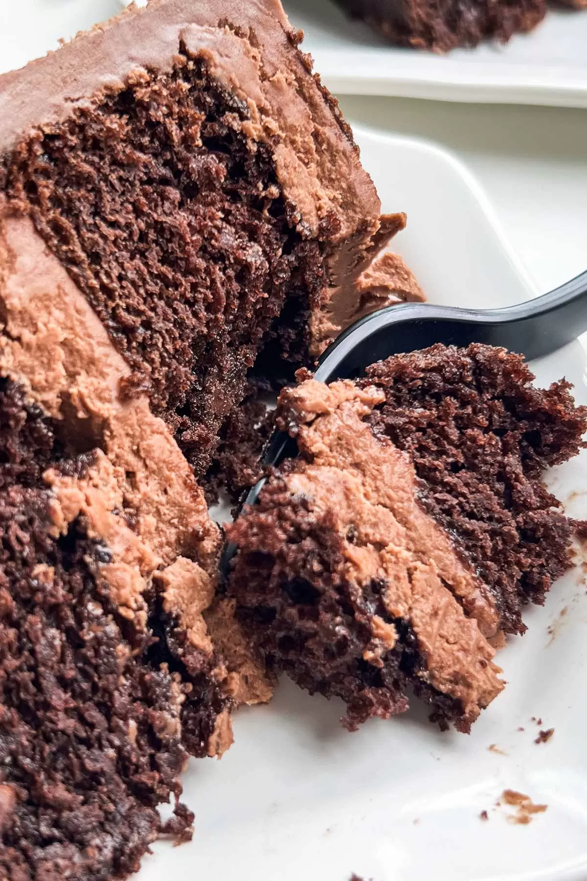 Moist slice of rich chocolate cake with a thick layer of chocolate buttercream, being cut into with a black plastic fork.