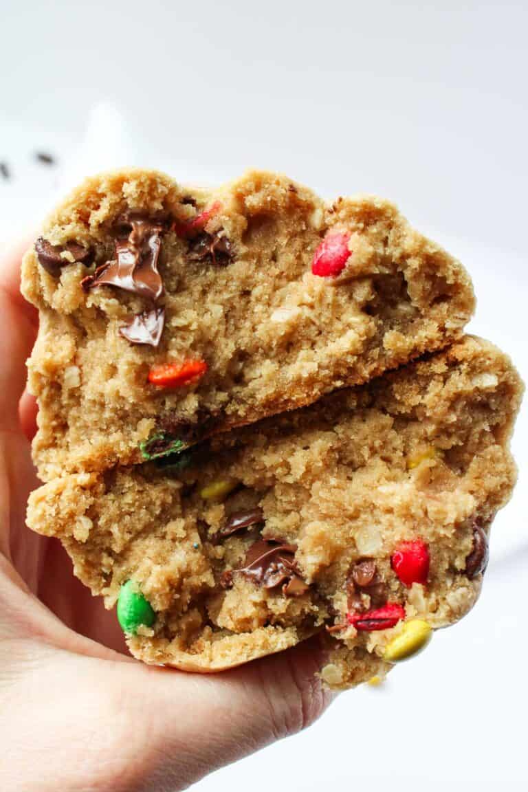 A close-up of a hand holding a freshly baked peanut butter cookie, revealing its soft, gooey center with colorful chocolate and candy pieces.